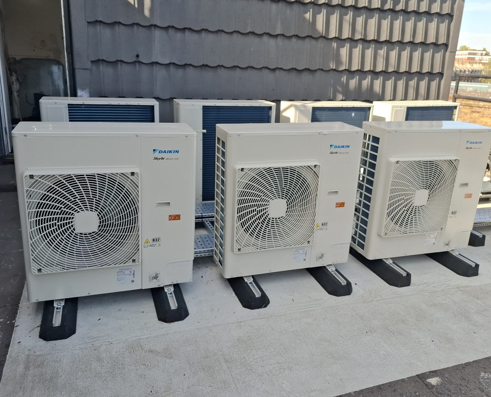 Rząd jednostek zewnętrznych klimatyzacji Daikin SkyAir na dachu, z widocznymi detalami montażu i szarą dachówką w tle. Widok z boku, lekko z góry.