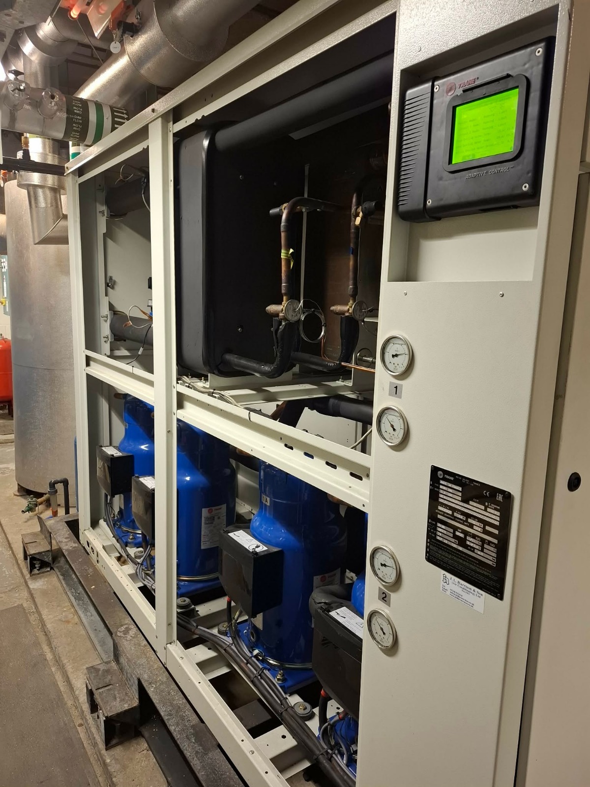 Agregat chłodniczy Trane z zielonym wyświetlaczem, manometrami i niebieskimi sprężarkami. Widoczne rury i elementy instalacji. Całość w metalowej obudowie.