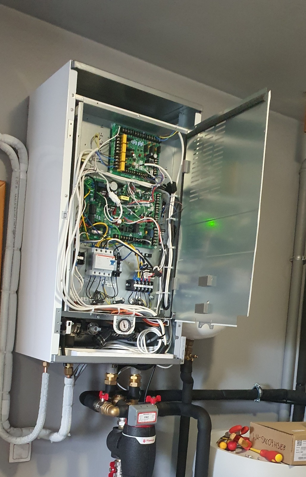Otwarty panel kotła grzewczego z widocznymi przewodami, elektroniką i rurami. Na dole filtry i zawory. Obok narzędzia i karton. Ujęcie od dołu.