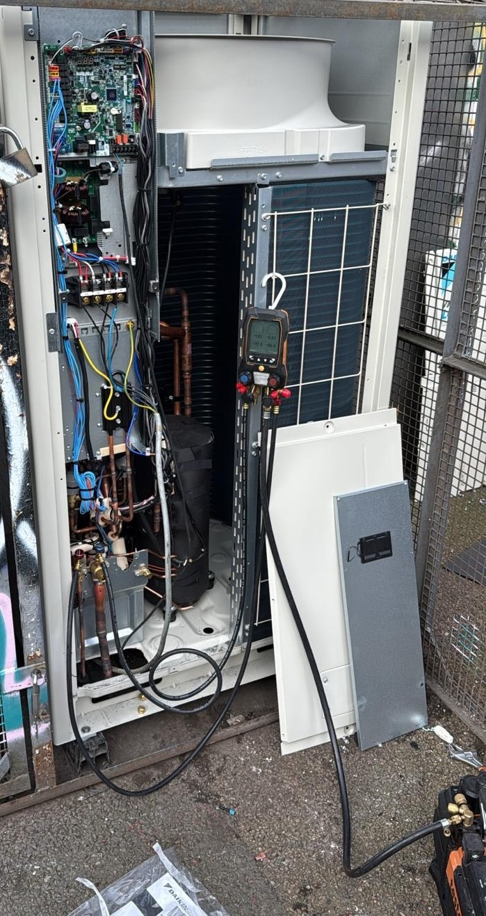 Otwarta obudowa klimatyzatora z widocznymi elementami wewnętrznymi, w tym rurami miedzianymi, elektroniką i narzędziami diagnostycznymi. Widoczne panele obudowy obok urządzenia.