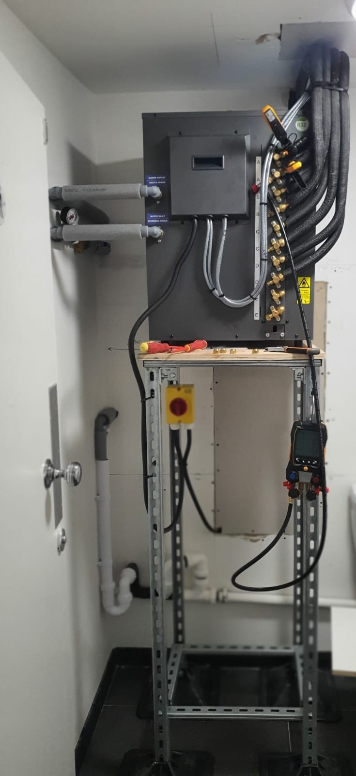 Szara centrala wentylacyjna z rurami, zaworami i narzędziami pomiarowymi na metalowym stojaku w pomieszczeniu. Widoczne przewody i elementy instalacji wodnej.