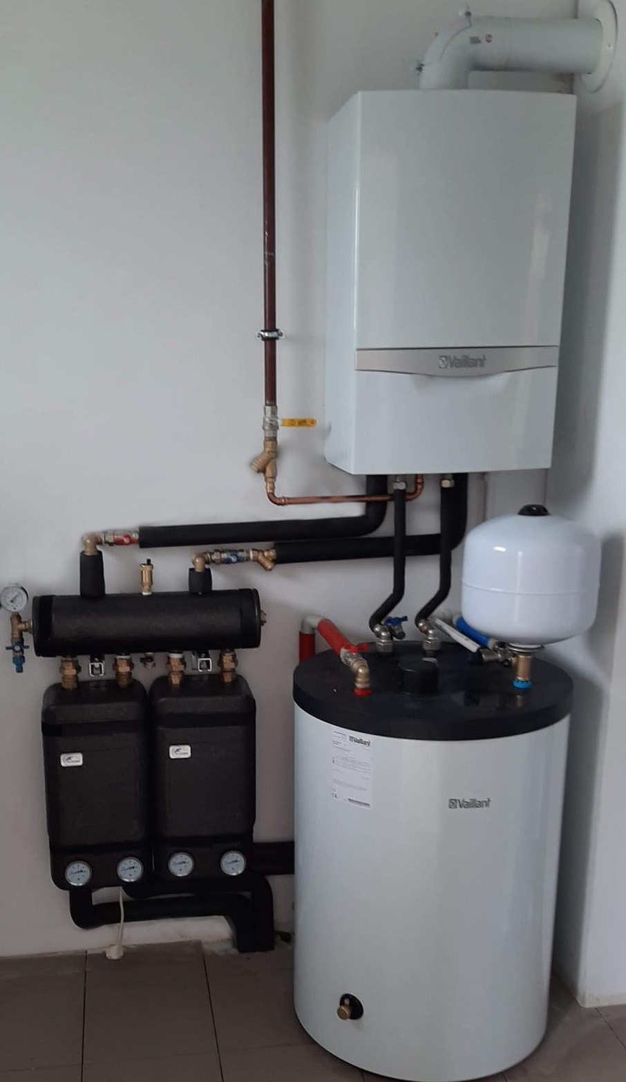 Zestaw instalacji grzewczej Vaillant z kotłem, zasobnikiem i grupą pompową, z widocznymi rurami miedzianymi i czarnymi, manometrami i zaworami, na tle białej ściany i szarej podłogi.