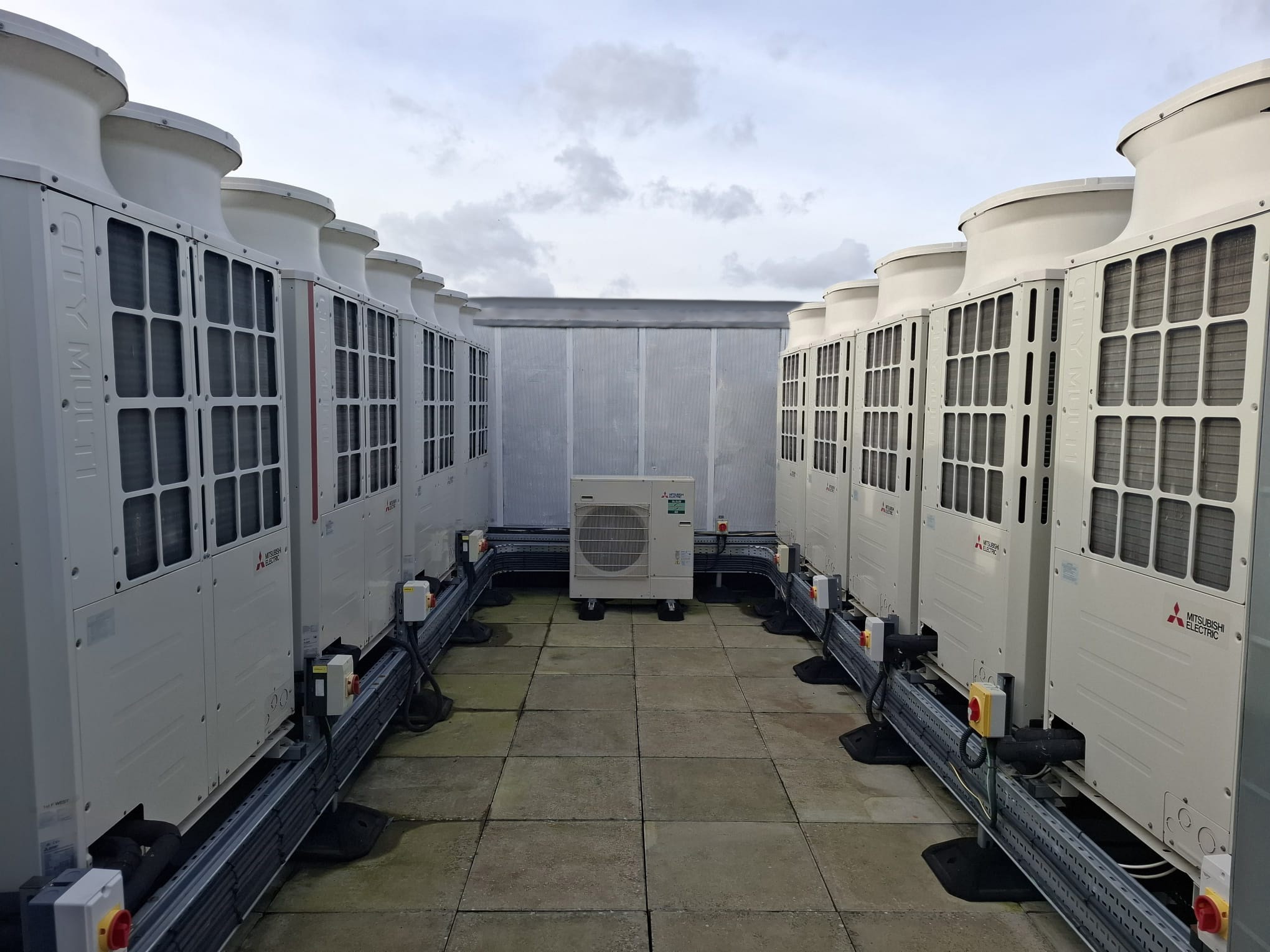 Rząd białych, przemysłowych klimatyzatorów Mitsubishi Electric na dachu budynku, z szarymi płytami chodnikowymi i pochmurnym niebem w tle. Widok z perspektywy.