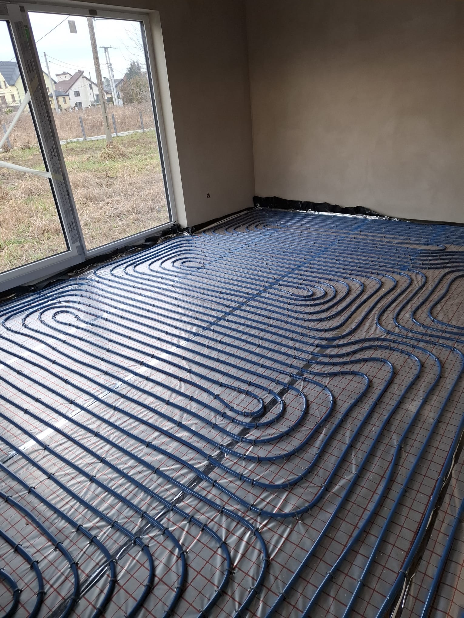 Niebieskie rury ogrzewania podłogowego ułożone spiralnie na folii izolacyjnej w surowym wnętrzu z dużym oknem. Widok na zewnątrz na domy i łąkę.