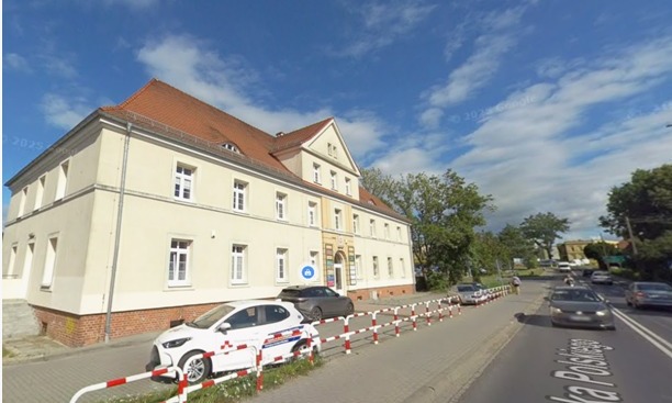 Elewacja dwupiętrowego budynku z czerwonym dachem, widziana z ulicy z zaparkowanymi samochodami i barierami ochronnymi. Błękitne niebo z chmurami w tle.