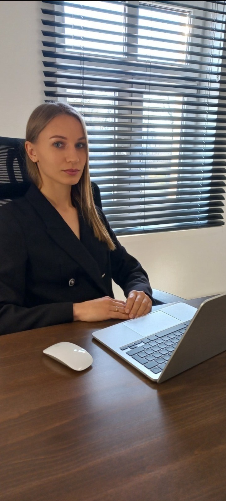 Kobieta w czarnej marynarce siedzi przy biurku z laptopem i myszką, w tle okno z żaluzjami. Profesjonalny wizerunek prawnika.