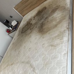 Royal clean - Zabrudzony materac na łóżku z drewnianą ramą. Widoczne plamy i przebarwienia na powierzchni materaca. Na szafce nocnej lampka i budzik.