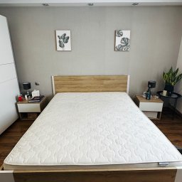 Royal clean - Czysty materac na łóżku z drewnianym zagłówkiem w sypialni. Widoczny fragment odkurzacza piorącego. Dwa stoliki nocne z lampkami i dekoracjami.