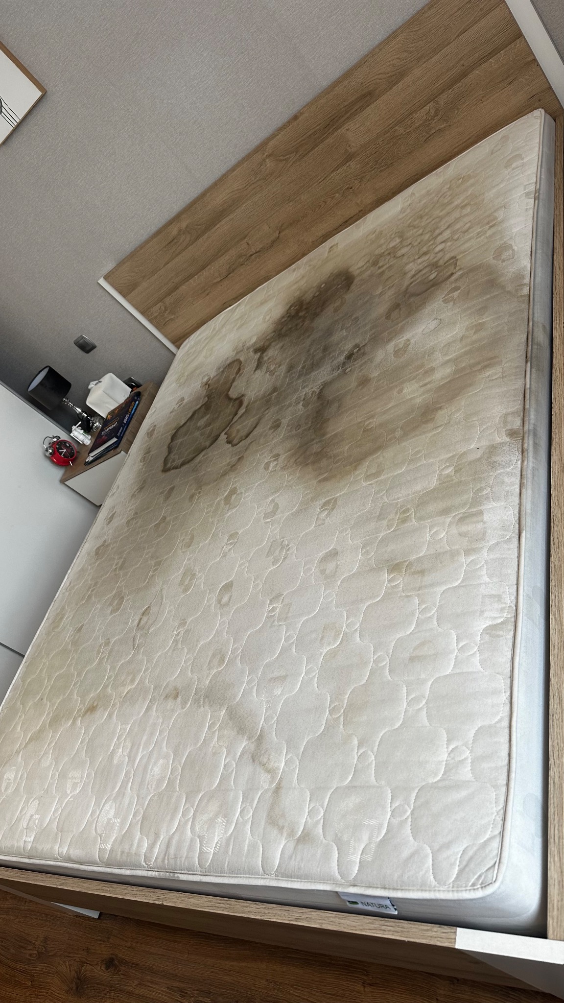 Zabrudzony materac na łóżku z drewnianą ramą. Widoczne plamy i przebarwienia na powierzchni materaca. Na szafce nocnej lampka i budzik.