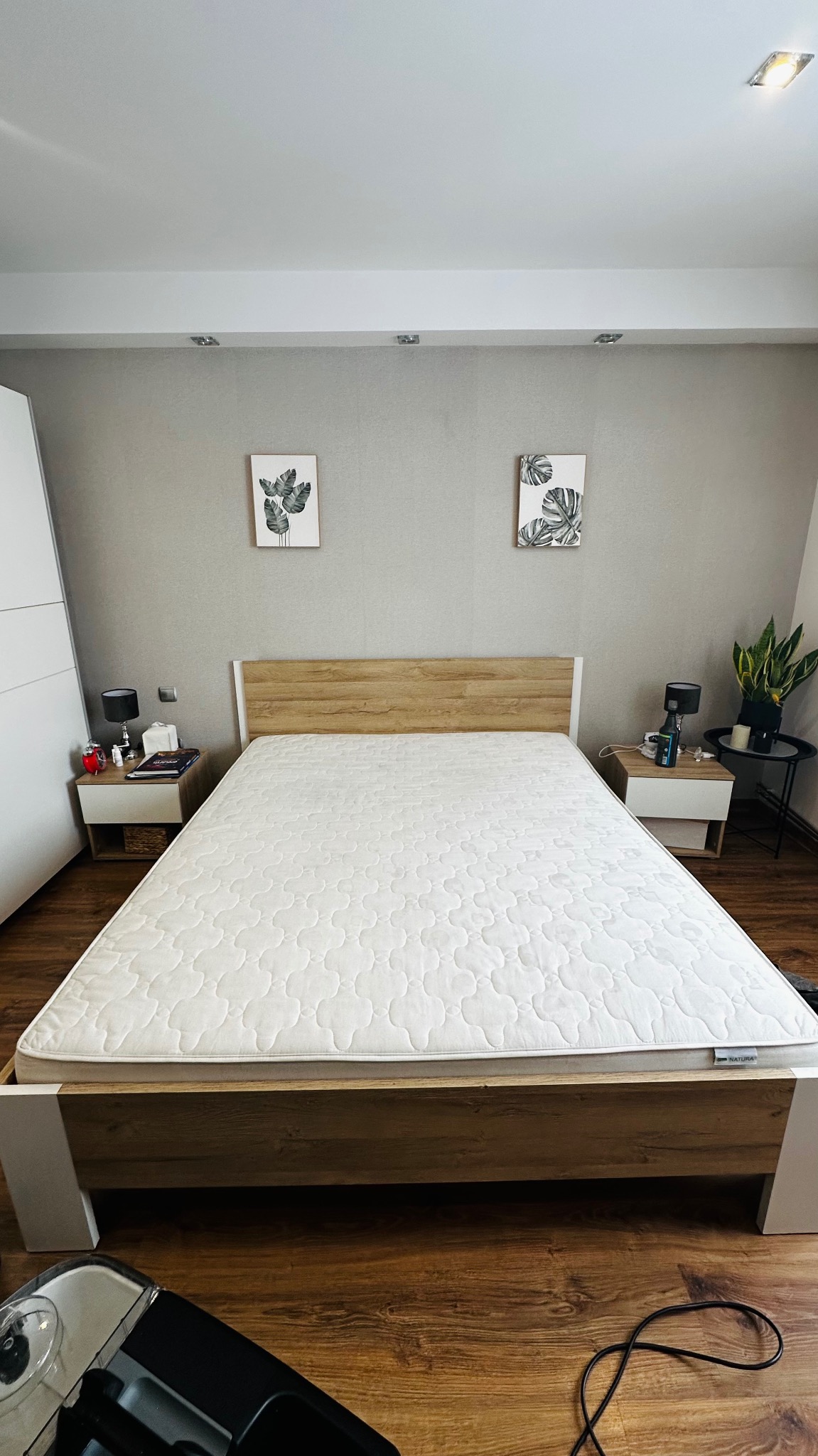 Czysty materac na łóżku z drewnianym zagłówkiem w sypialni. Widoczny fragment odkurzacza piorącego. Dwa stoliki nocne z lampkami i dekoracjami.