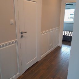 Rafoss Rafał Ossowski - Białe panele ścienne z dekoracyjnymi ramkami w korytarzu, brązowe płytki podłogowe, białe drzwi i widok na pokój z łóżkiem. Subtelne oświetlenie.