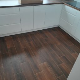 Rafoss Rafał Ossowski - Nowoczesna kuchnia z białymi, lakierowanymi frontami szafek i ciemnobrązową podłogą imitującą drewno. Widoczny fragment blatu i płyty indukcyjnej.