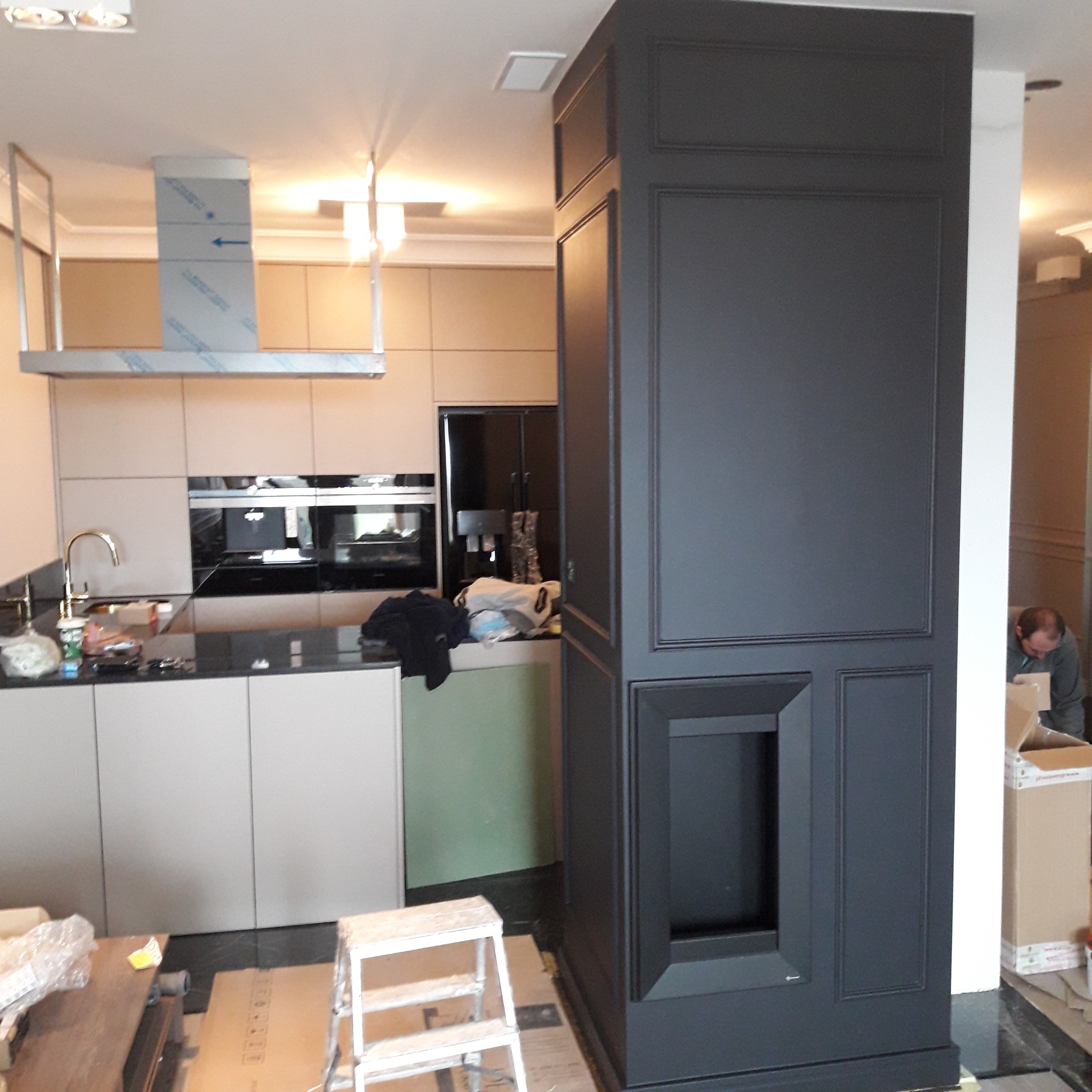 Ciemnoszara zabudowa meblowa w kuchni z beżowymi szafkami, widoczny fragment drabiny i osoba w tle, sugerująca prace wykończeniowe.