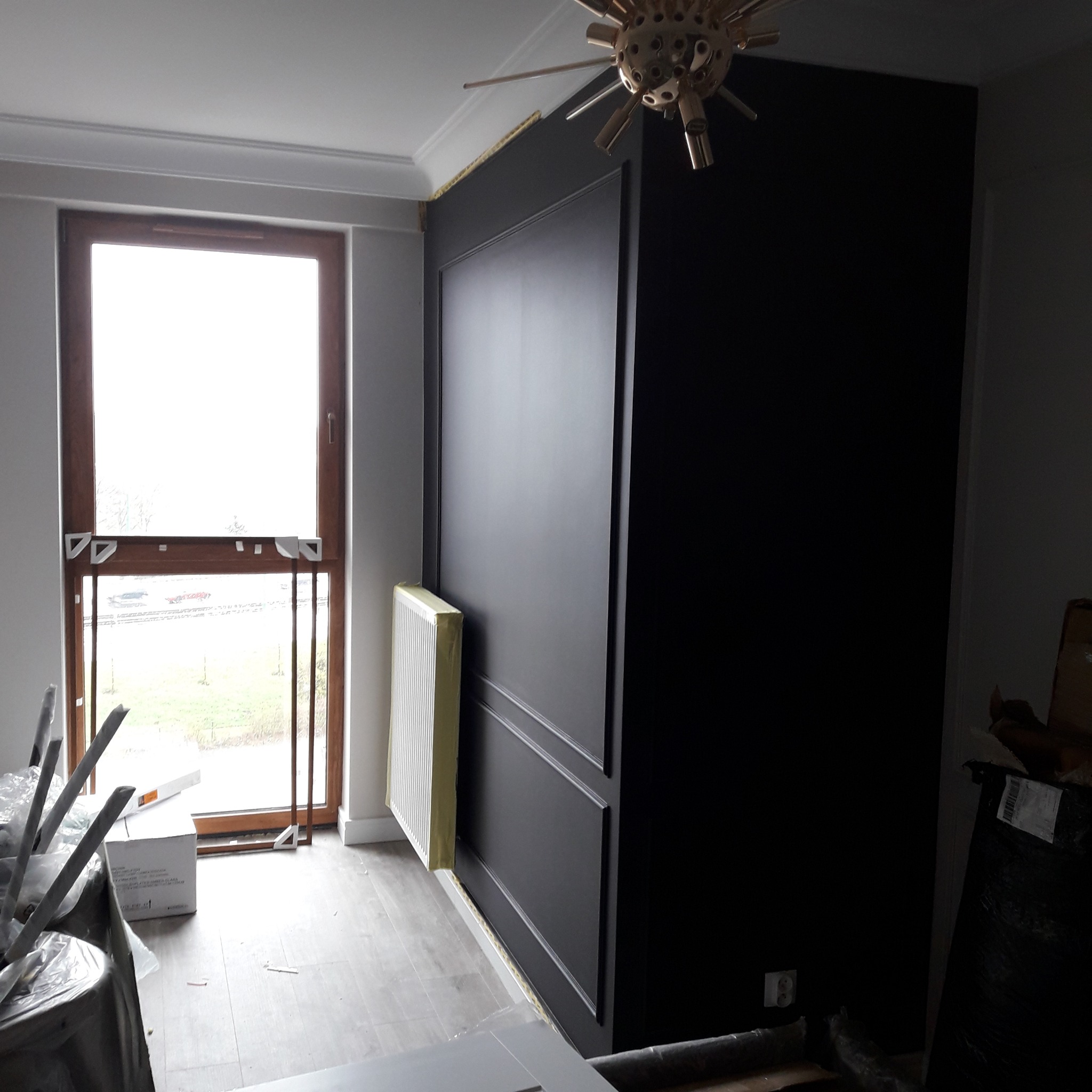 Ciemna ściana z dekoracyjnymi panelami w trakcie prac wykończeniowych, widoczny grzejnik zabezpieczony folią i elementy wyposażenia wnętrza.