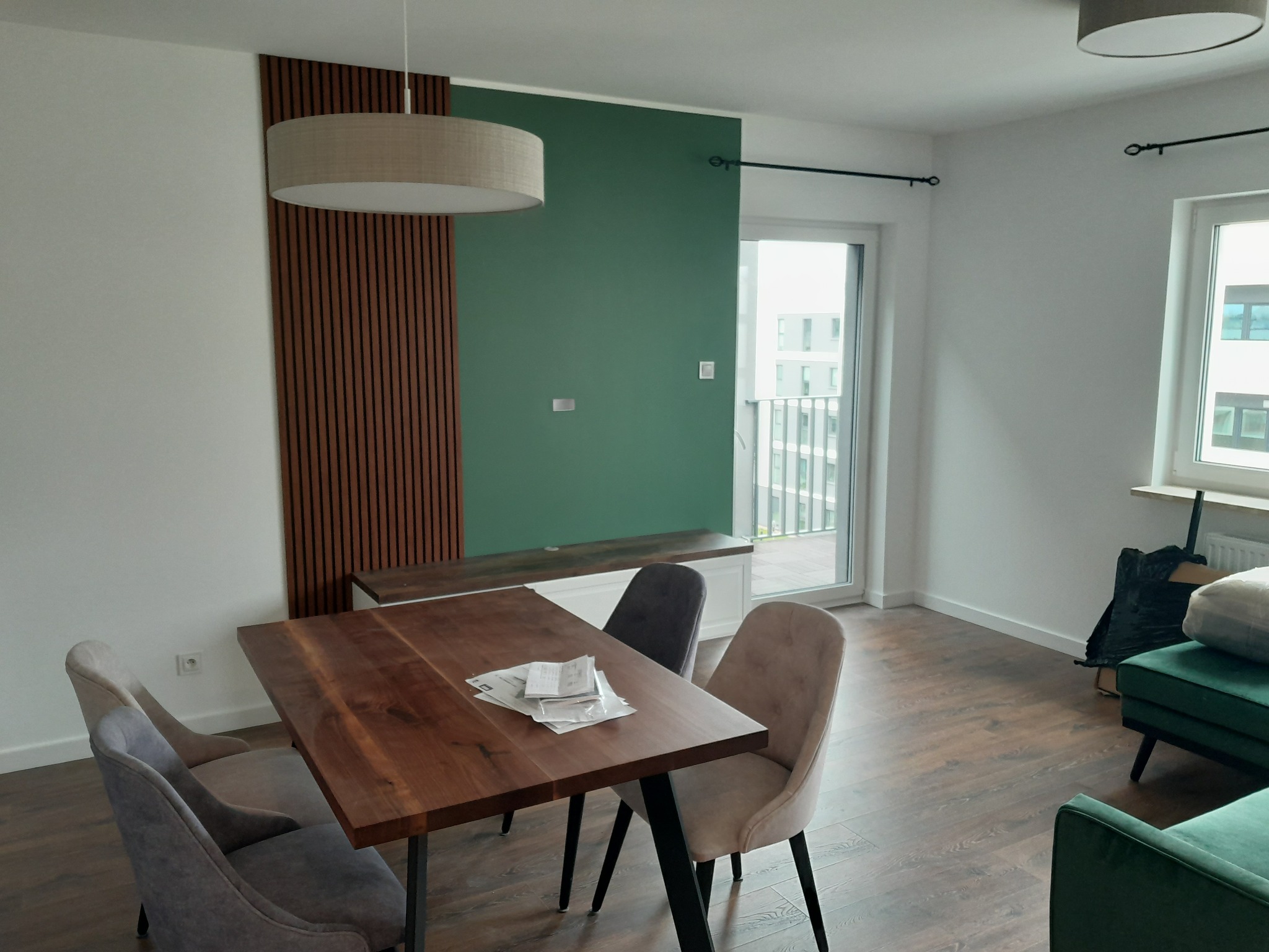 Nowoczesne wnętrze: drewniany stół z krzesłami, zielona ściana z dekoracyjnymi panelami, lampa wisząca. Minimalistyczny design, jasne pomieszczenie z dużym oknem.