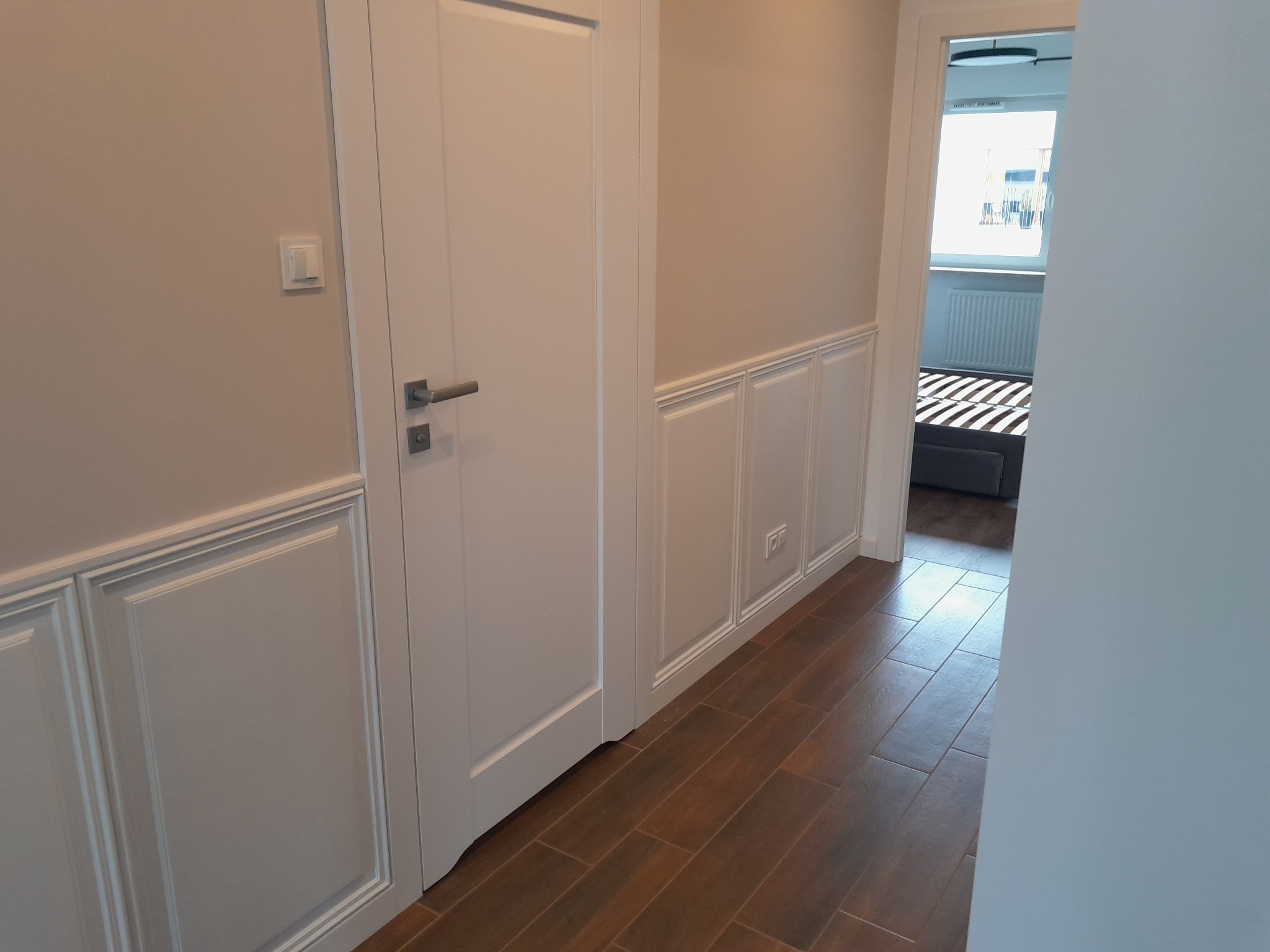 Białe panele ścienne z dekoracyjnymi ramkami w korytarzu, brązowe płytki podłogowe, białe drzwi i widok na pokój z łóżkiem. Subtelne oświetlenie.