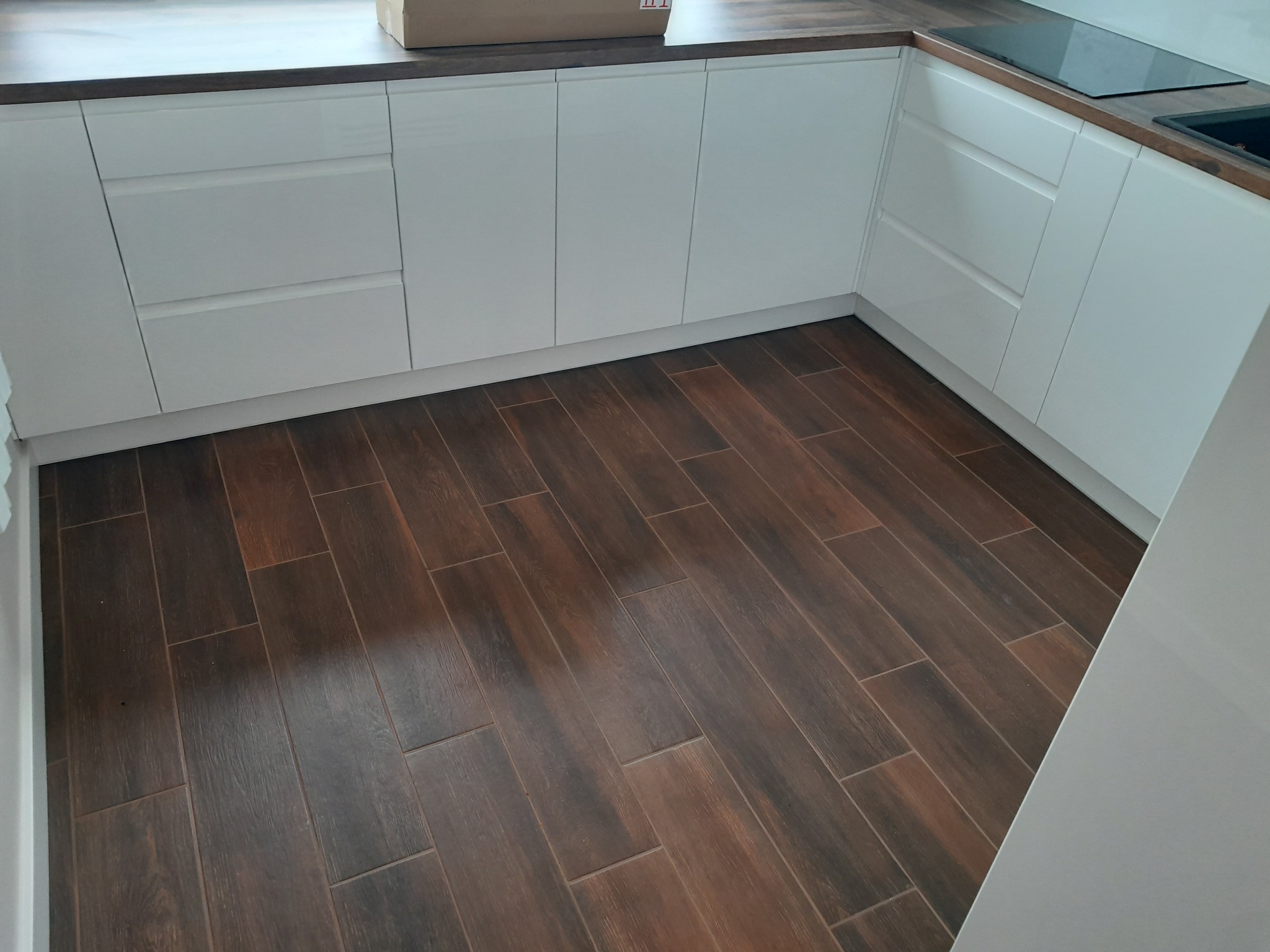 Nowoczesna kuchnia z białymi, lakierowanymi frontami szafek i ciemnobrązową podłogą imitującą drewno. Widoczny fragment blatu i płyty indukcyjnej.