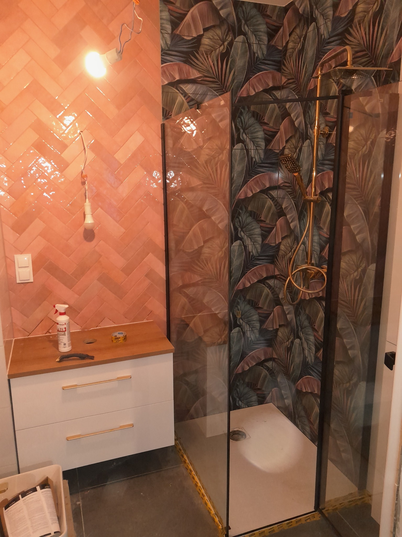 Wykończona łazienka z kabiną prysznicową, umywalką z szafką i oryginalnymi płytkami w odcieniach różu i zieleni z motywem liści. Widoczne narzędzia i materiały.