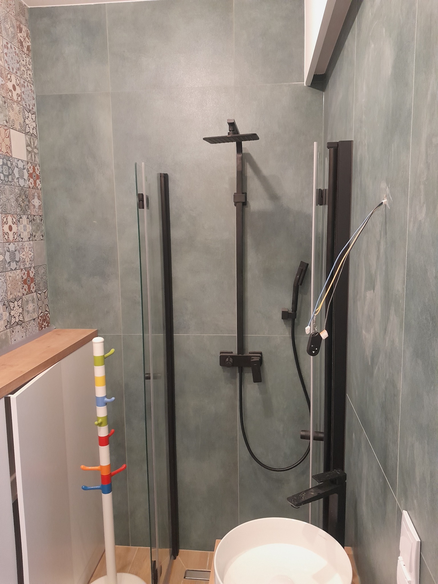 Nowoczesna łazienka z kabiną prysznicową typu walk-in, czarną armaturą i umywalką. Widoczne wystające przewody elektryczne. Na ścianach szaro-zielone płytki.