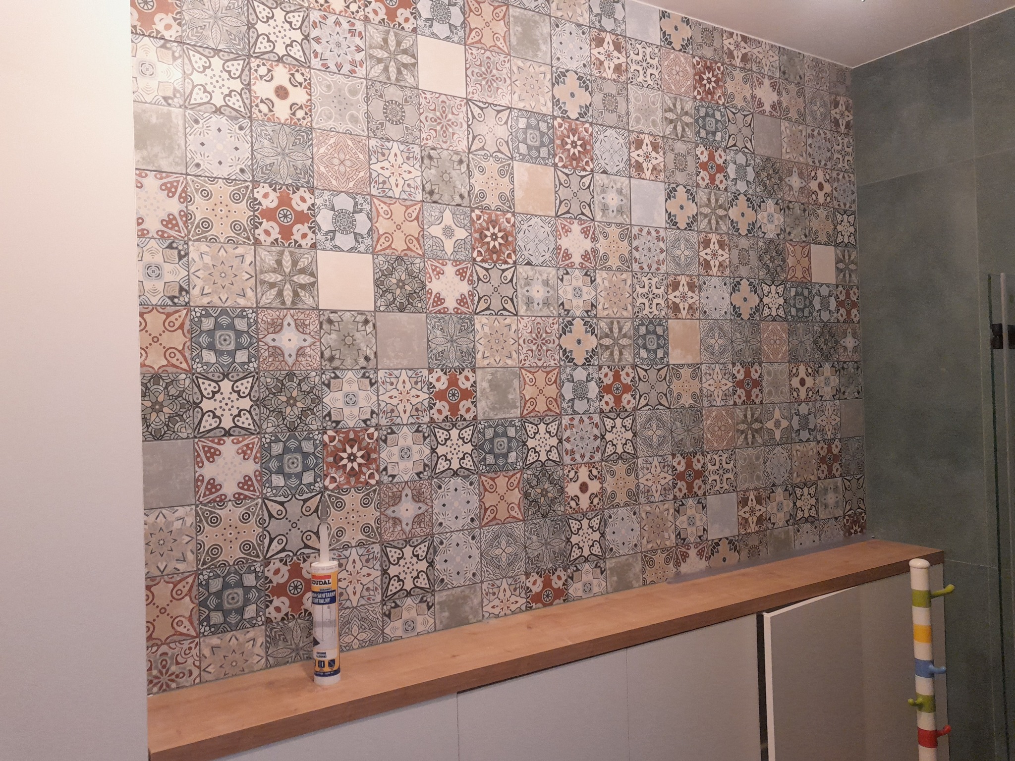 Ściana wyłożona dekoracyjnymi, wzorzystymi płytkami w łazience. Na blacie tuba silikonu sanitarnego. Widoczny fragment szafki i wieszak na ręczniki.