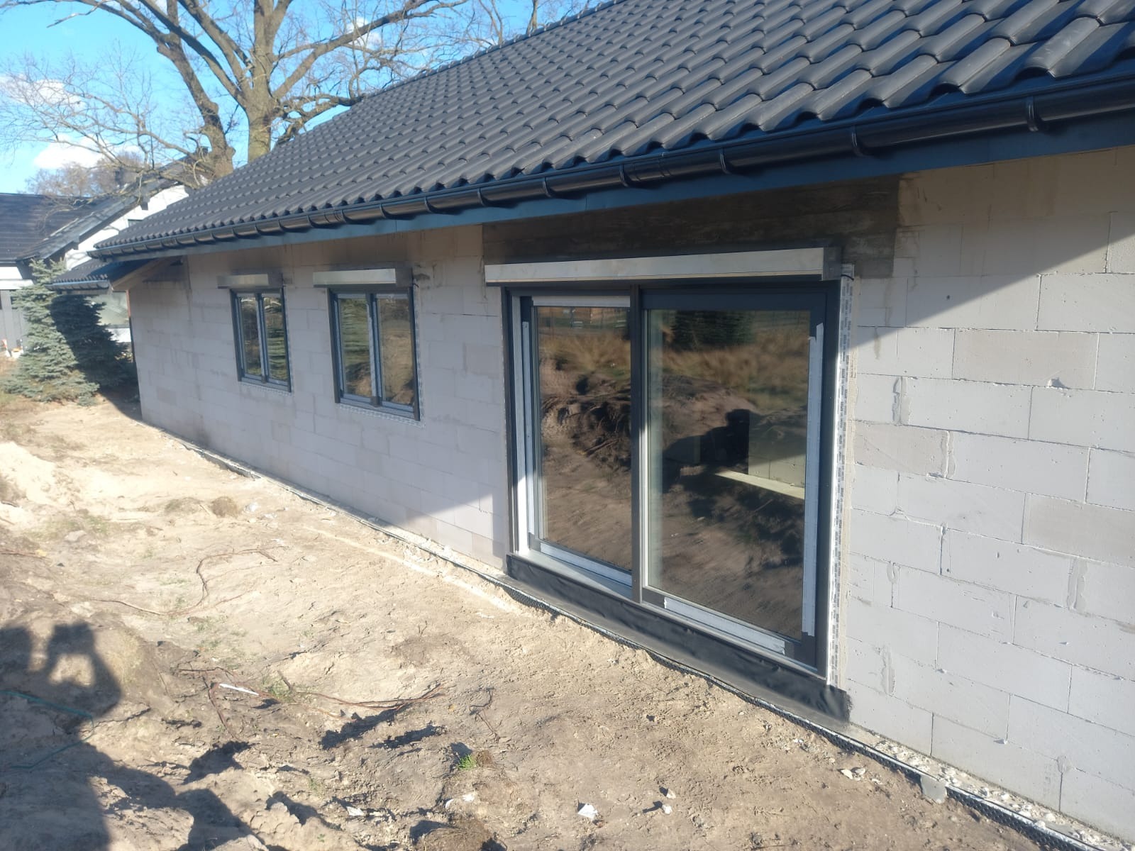 Szare okna i drzwi przesuwne w surowym budynku z betonu komórkowego. Widoczne rolety zwinięte nad oknami. W tle drzewo bez liści i fragment dachu sąsiedniego domu.