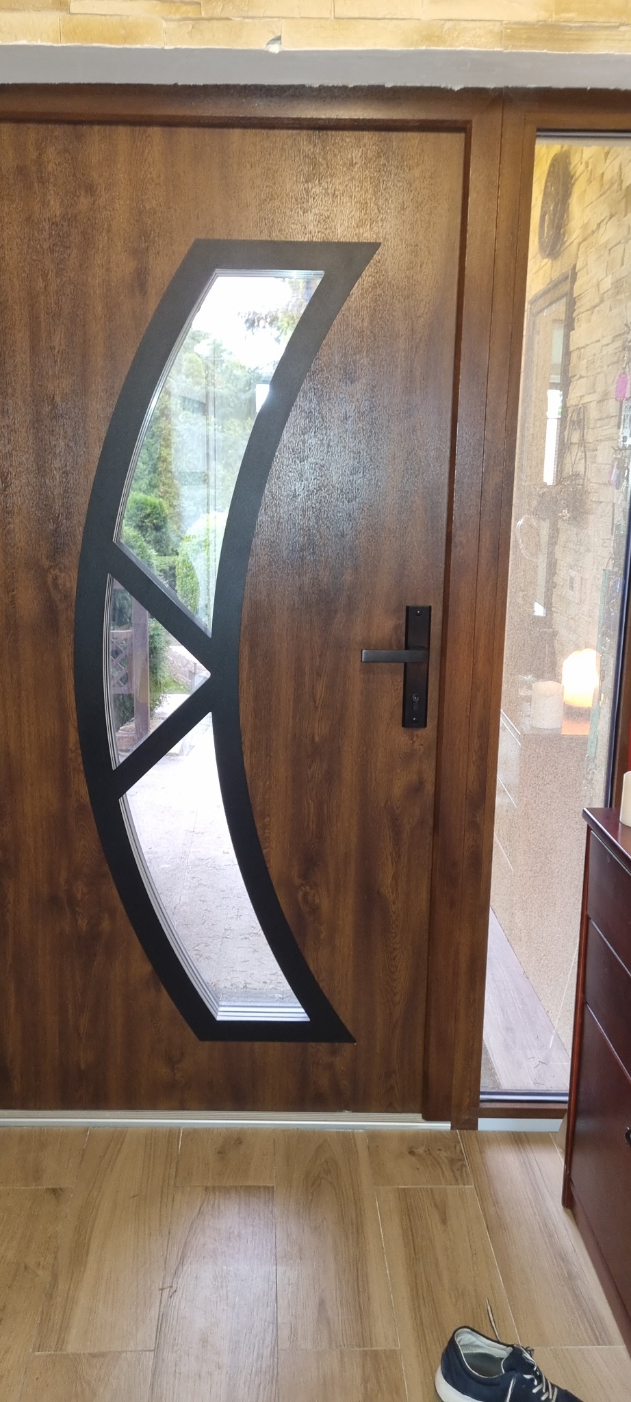 Brązowe drzwi zewnętrzne z oryginalnym, półksiężycowatym przeszkleniem i czarną klamką, widoczne od strony wnętrza, z fragmentem podłogi i buta.