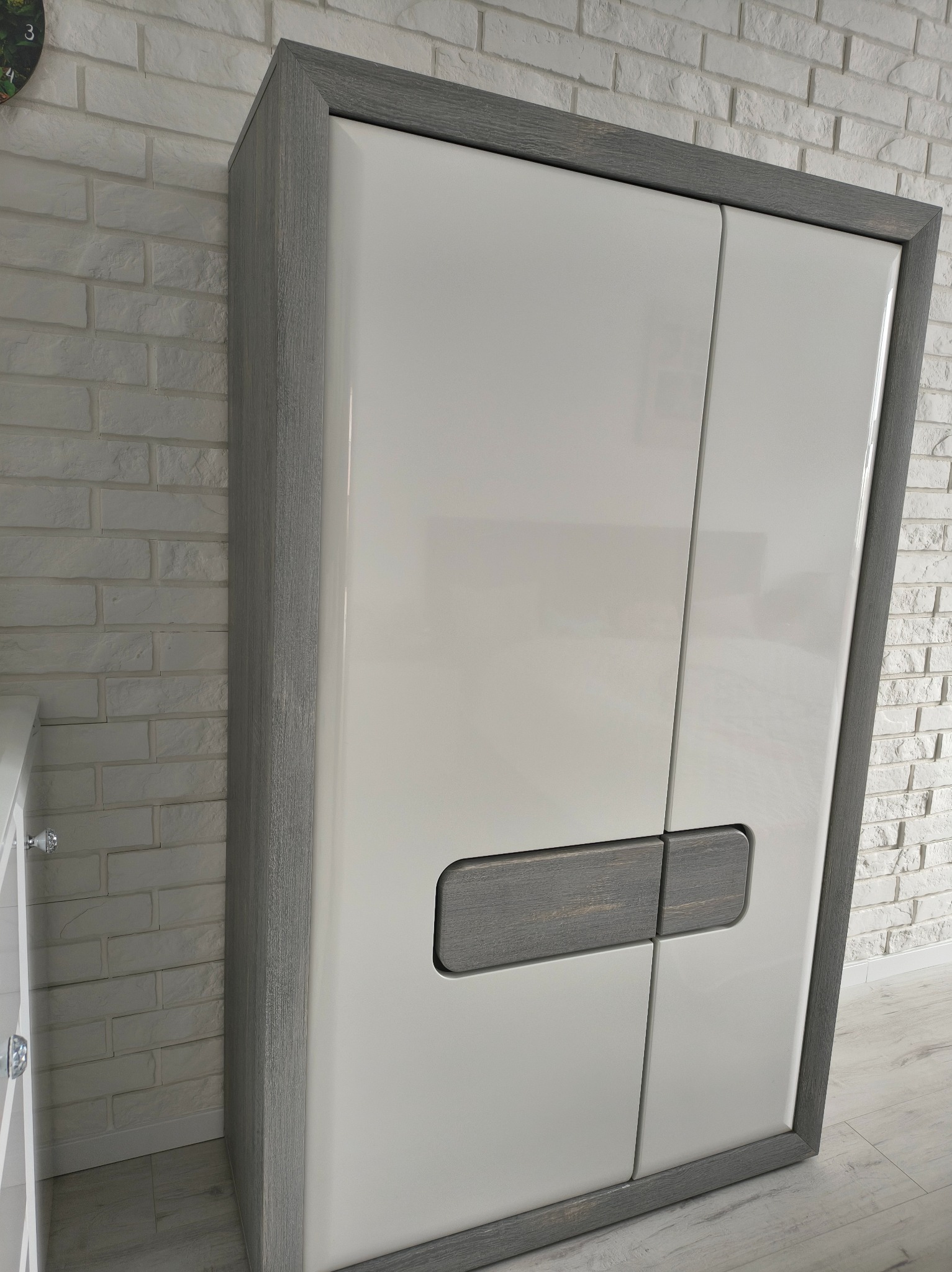 Nowoczesna, dwudrzwiowa szafa z białymi frontami i szarą ramą, ustawiona na tle ściany z białej cegły. Widoczny fragment podłogi z jasnymi panelami.