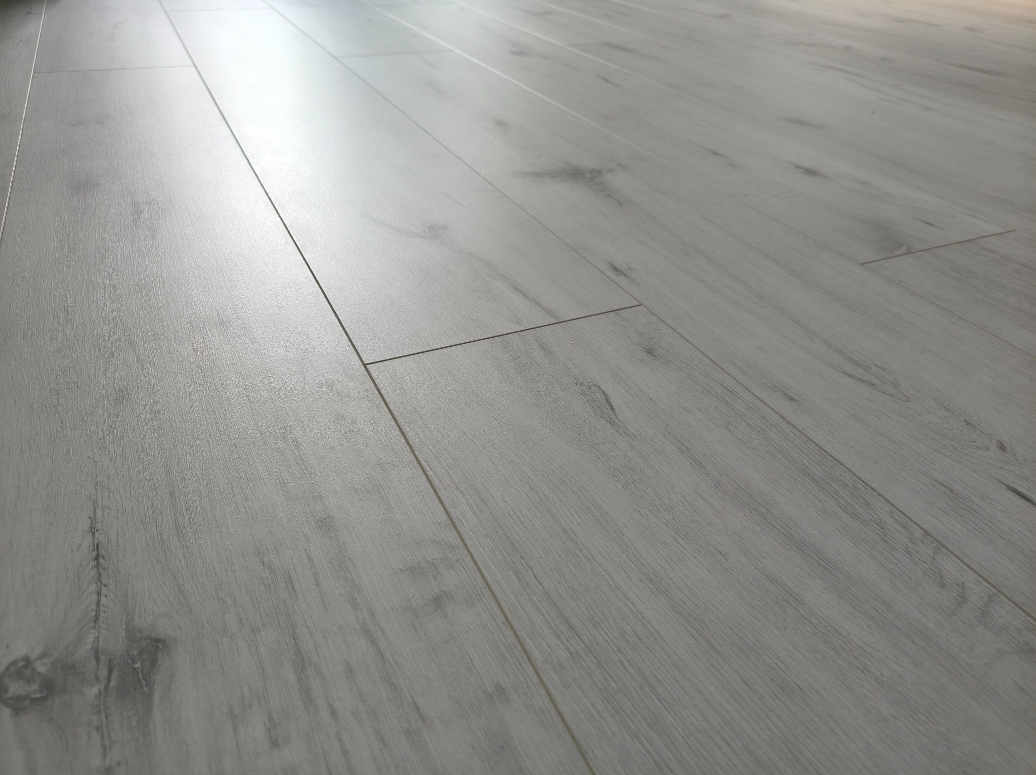 Jasnoszara podłoga z paneli laminowanych, ułożona pod kątem, z widoczną strukturą drewna i delikatnym połyskiem, ukazująca precyzję łączeń.