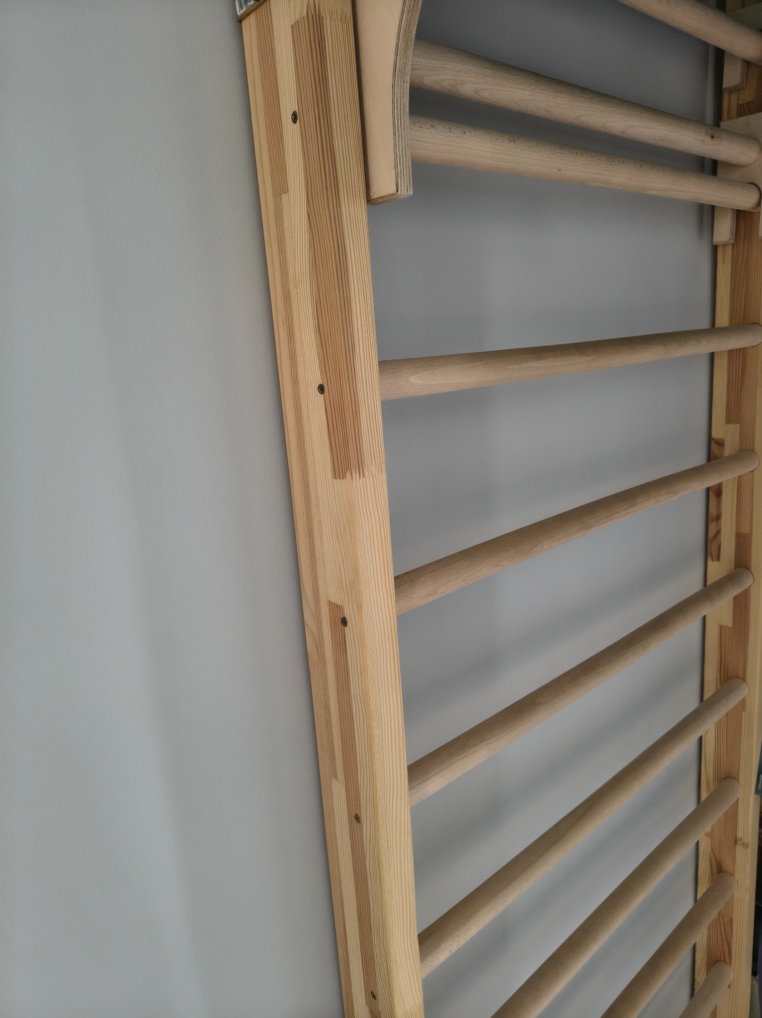 Drewniana drabinka gimnastyczna przymocowana do szarej ściany. Widoczne naturalne usłojenie drewna i metalowe śruby mocujące. Ujęcie z boku, oświetlenie naturalne.
