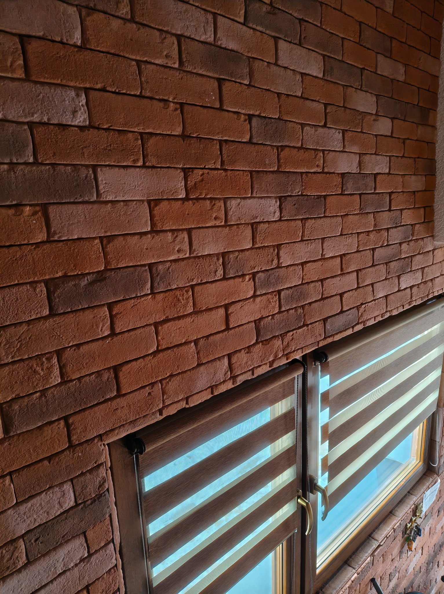 Ściana z cegły klinkierowej w odcieniach czerwieni i brązu, z widocznymi oknami przesłoniętymi roletami w stylu zebra, tworzącymi ciepły, rustykalny klimat.