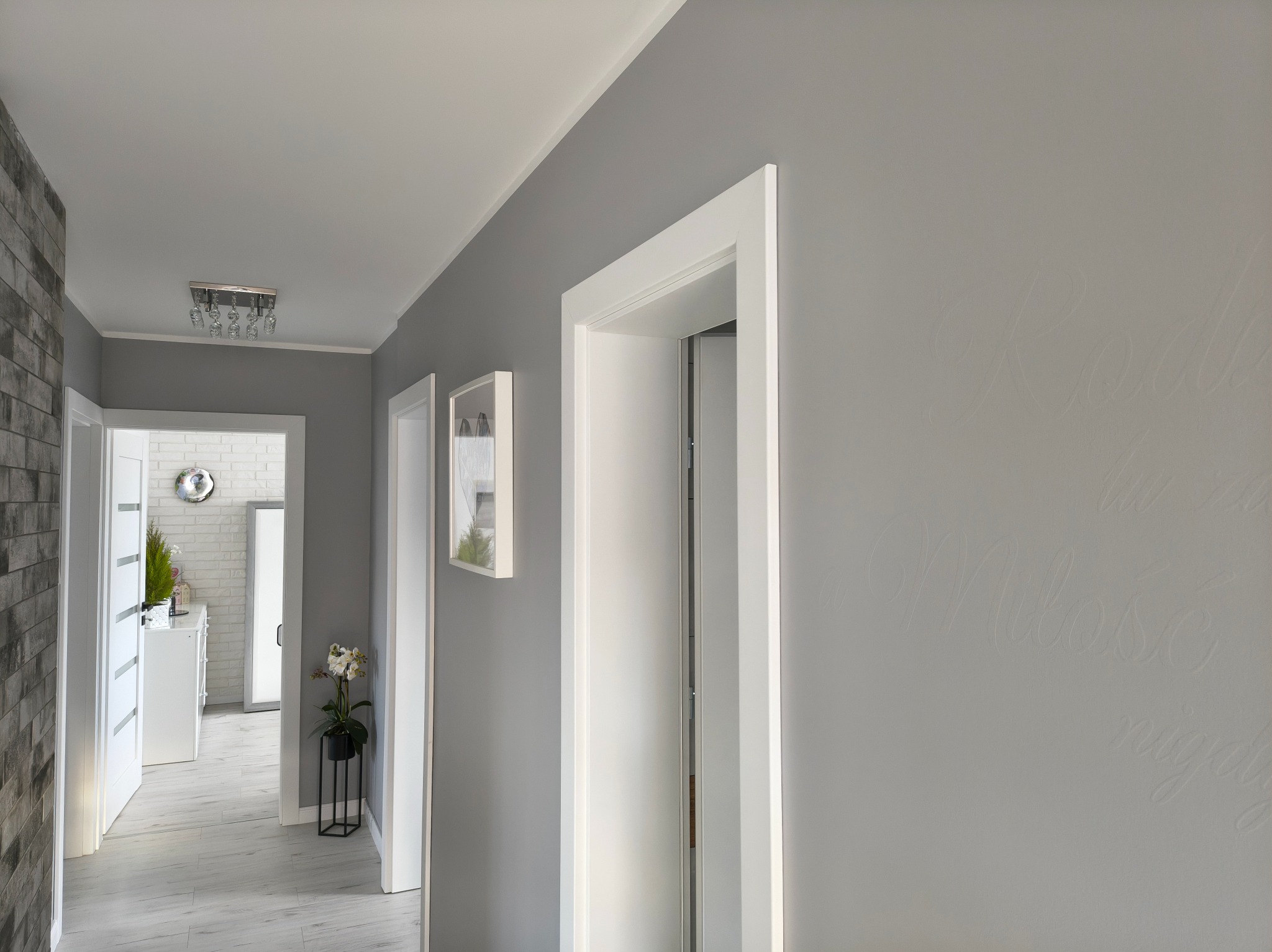 Jasnoszary korytarz po malowaniu z białymi drzwiami i dekoracyjnym napisem na ścianie. Widok na wnętrze z kwiatami i ceglaną ścianą w tle.