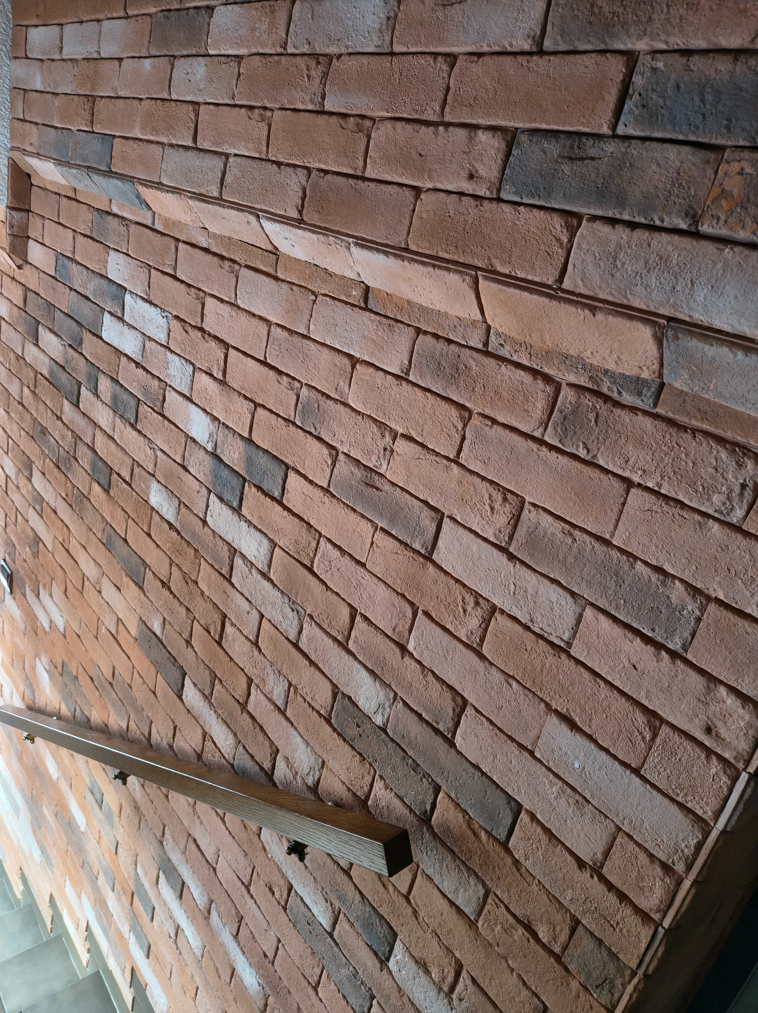 Ściana wykończona czerwoną cegłą klinkierową z drewnianą poręczą schodów. Widoczny fragment schodów. Nietypowe ułożenie cegieł pod kątem.
