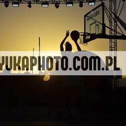 YUKA PHOTO&PR - Silueta koszykarza oddającego rzut do kosza na tle zachodzącego słońca. W tle widoczne maszty jachtów i konstrukcja kosza z napisem Heraklion.