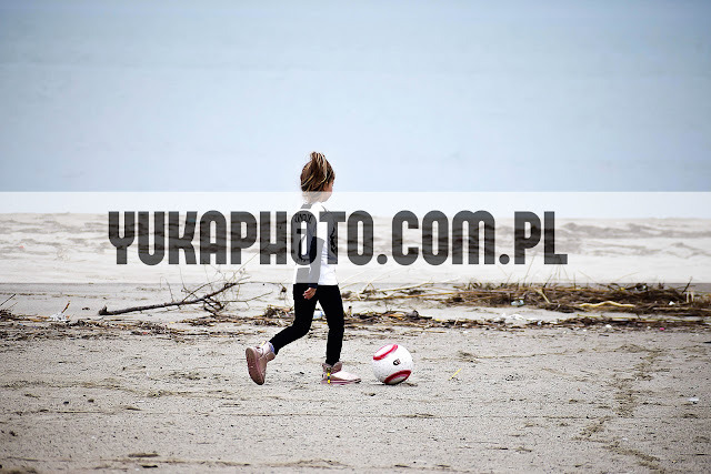 Dziewczynka w czarnych legginsach i jasnej bluzce kopie piłkę na piaszczystej plaży. Ujęcie z boku, w tle widać morze i niebo. Na piasku leżą gałęzie.