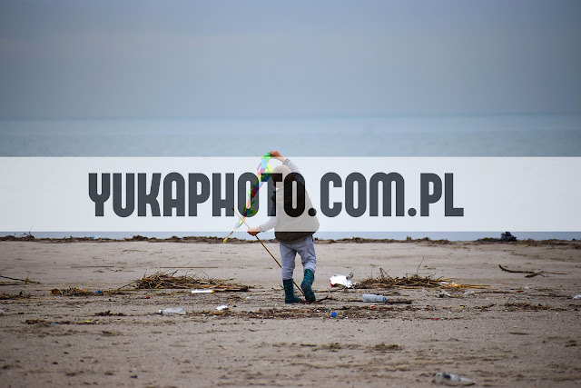 Chłopiec w kaloszach, tyłem, trzyma latawiec na plaży z widokiem na morze. Plaża zaśmiecona, pochmurne niebo. Znak wodny YUKAPHOTO.COM.PL.