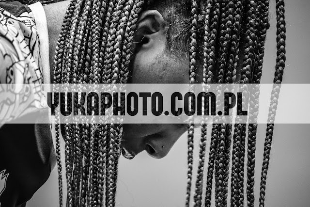 Czarno-białe zbliżenie na twarz kobiety z zaplecionymi warkoczykami, widoczny piercing w nosie. Ujęcie z góry, w tle jasna ściana. Na zdjęciu widoczny znak wodny.