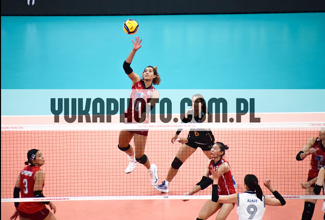 Siatkarka w wyskoku atakuje piłkę podczas meczu. Widoczne siatka, tło w kolorze turkusowym i inne zawodniczki. Dynamiczne ujęcie z akcji sportowej.