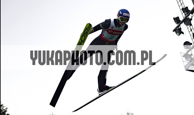 Skoczek narciarski w locie na tle jasnego nieba, z widocznymi nartami i kombinezonem. W tle oświetlenie skoczni. Zdjęcie z widocznym znakiem wodnym.