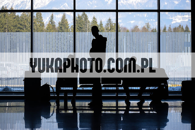 Sylwetka mężczyzny w poczekalni na tle okna z widokiem na góry. Bagaże obok krzeseł, refleksy na podłodze. Minimalistyczna kompozycja.