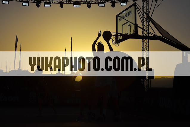 Silueta koszykarza oddającego rzut do kosza na tle zachodzącego słońca. W tle widoczne maszty jachtów i konstrukcja kosza z napisem Heraklion.