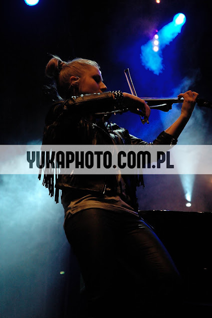 Kobieta w skórzanej kurtce z frędzlami gra na elektrycznych skrzypcach na scenie oświetlonej niebieskimi światłami. Ujęcie z dołu, widoczny znak wodny.