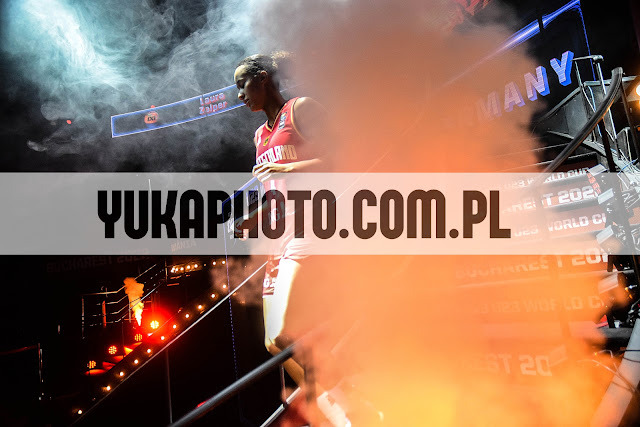 Kobieta w sportowym stroju wchodzi po schodach w dymie i świetle reflektorów podczas wydarzenia sportowego. Widoczne logo i napisy związane z zawodami.