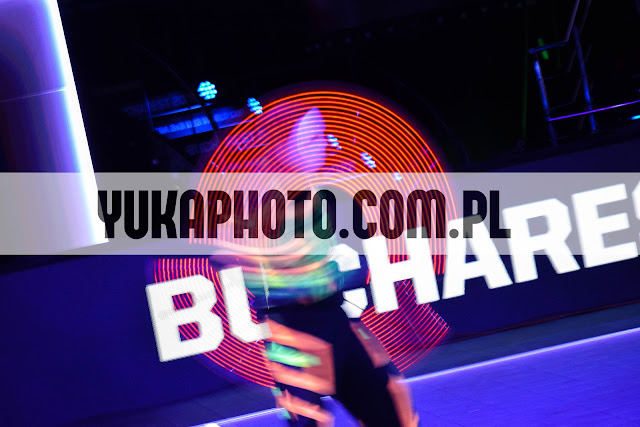 Rozmazane światła tworzą okrągły wzór na ciemnym tle, z widocznym napisem 'YUKAPHOTO.COM.PL' i 'BUCHAREST'. Dynamiczne ujęcie z efektem długiej ekspozycji.