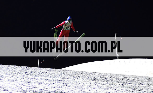 Skoczek narciarski w locie na tle czarnego nieba, widoczny numer 49 na kombinezonie, śnieżny zeskok. Dynamiczne ujęcie z zawodów.