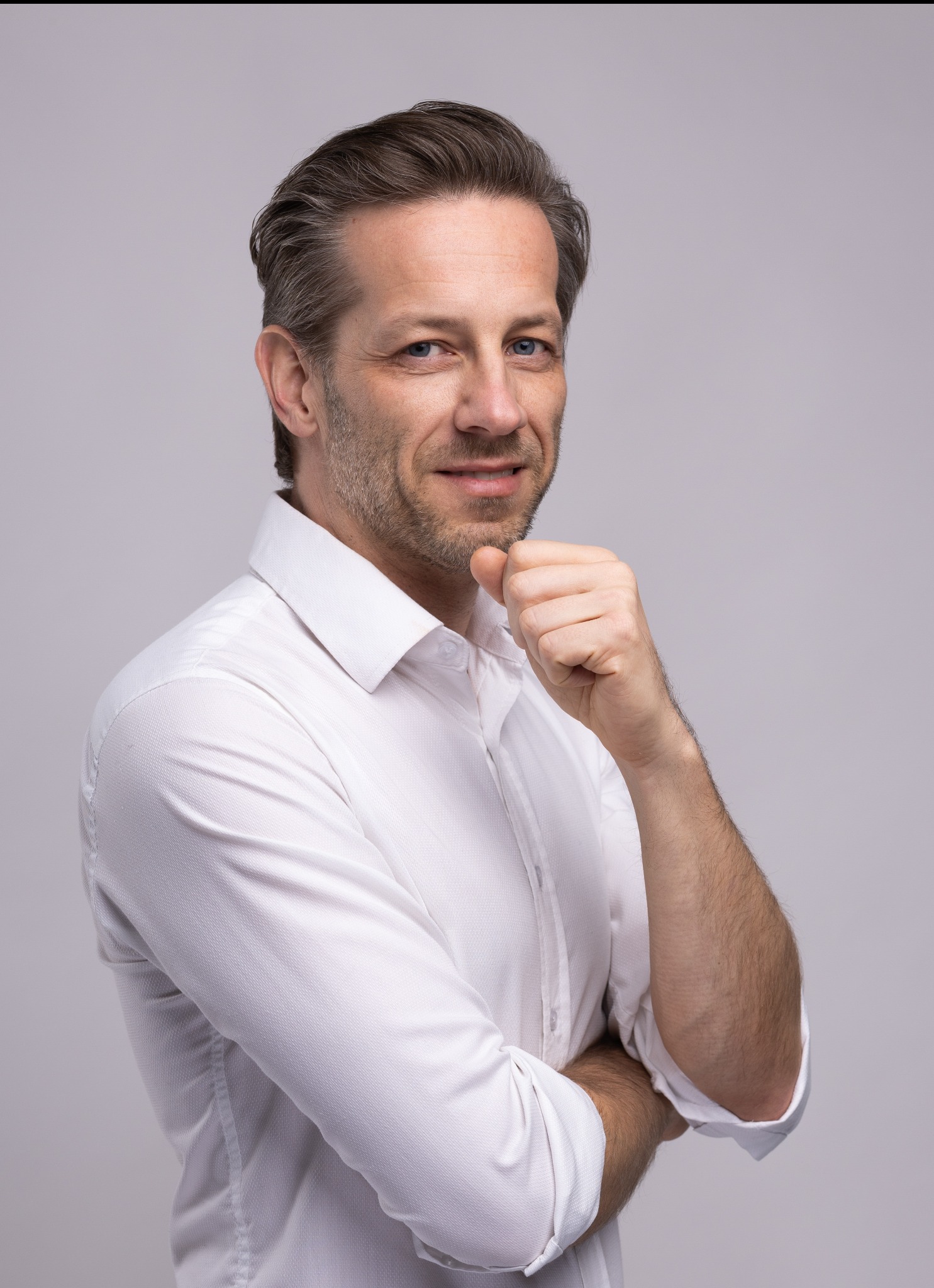 Portret mężczyzny w białej koszuli, z ręką pod brodą, na jasnym tle. Uśmiechnięty, z krótką brodą i niebieskimi oczami. Profesjonalny wizerunek.