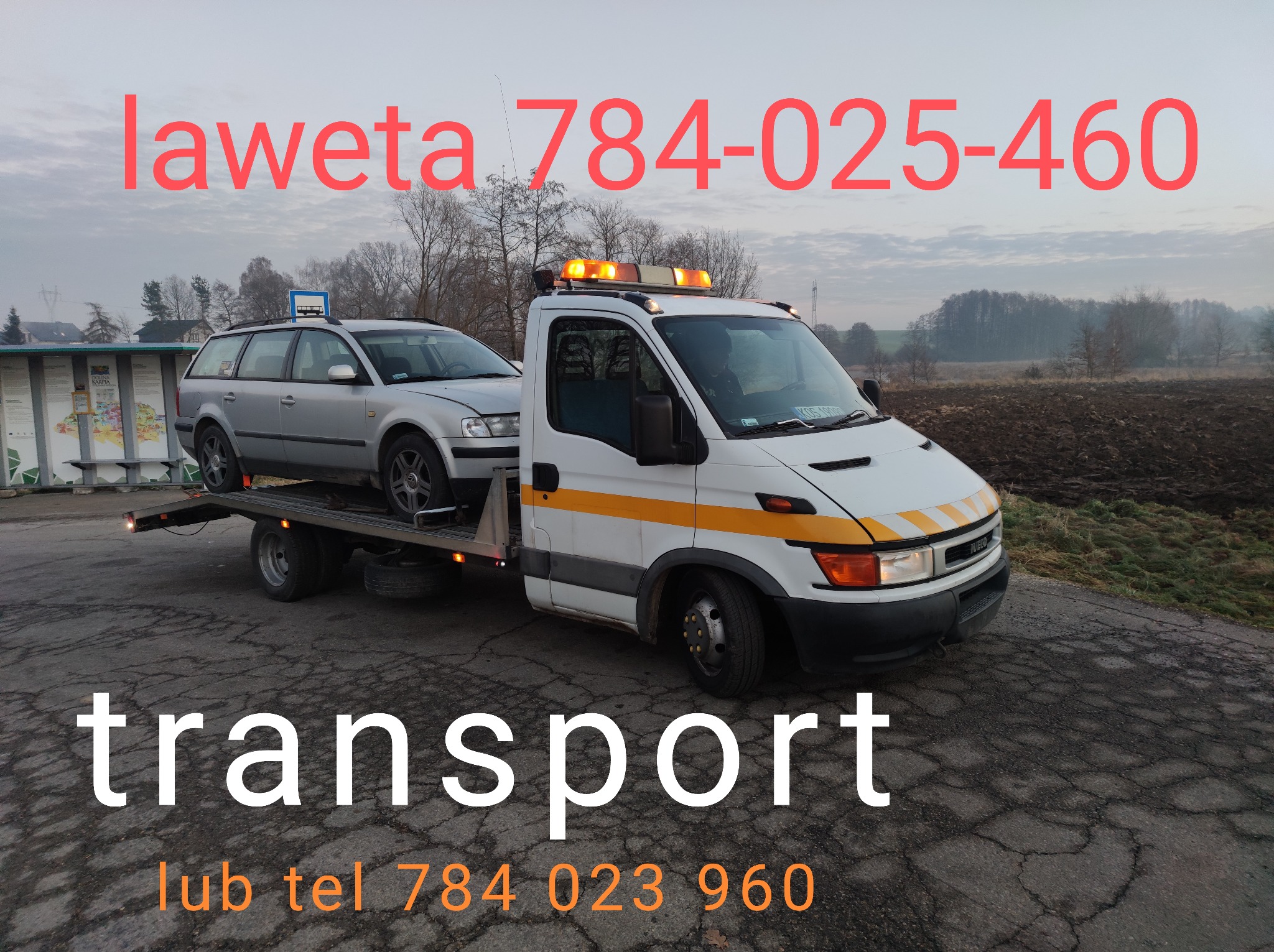 Laweta z srebrnym samochodem osobowym na platformie, widoczne numery telefonów kontaktowych na zdjęciu. Transport pojazdów na drodze.