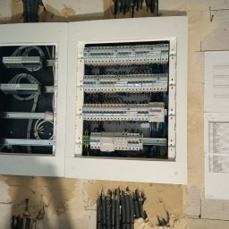 KOLBE - Otwarta, biała skrzynka rozdzielcza z widocznymi kablami i bezpiecznikami, zamontowana na surowej ścianie. Obok dokumentacja techniczna. Widoczne przewody w ścianie.