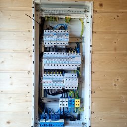 KOLBE - Rozdzielnica elektryczna Hager w obudowie, zamontowana na ścianie z boazerii. Widoczne zabezpieczenia, przewody w kolorach: niebieskim, czarnym, żółto-zielonym.