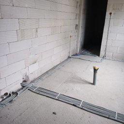 KOLBE - Szare rury w peszlu ułożone na surowym betonie podłogi w nowym budynku z białego pustaka. Część instalacji poprowadzona po ścianie. Widoczne otwory na gniazdka.