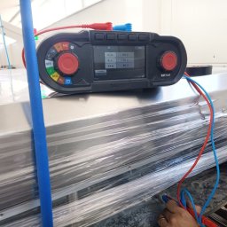 KOLBE - Miernik wielofunkcyjny Megger MFT-X1 z podłączonymi przewodami pomiarowymi, wyświetlający wyniki pomiarów napięcia. Urządzenie leży na metalowej powierzchni.